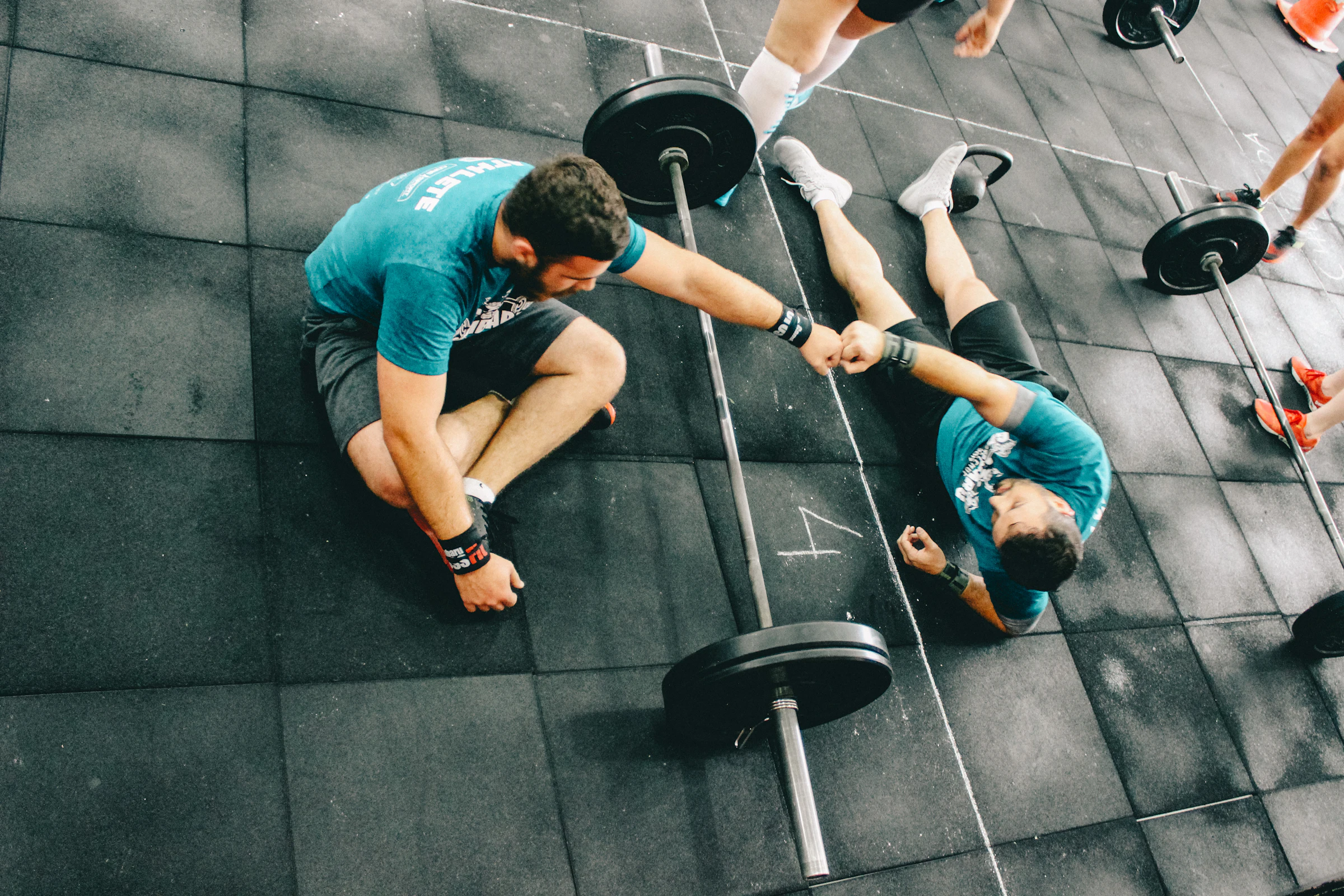 Dos atletas tendidos en el piso después de un WOD se chocan los puños junto a sus barras.